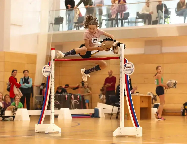Hobby Horsing-deelnemer springt met een Hobby Horse over een rode paal tijdens een indoor evenement