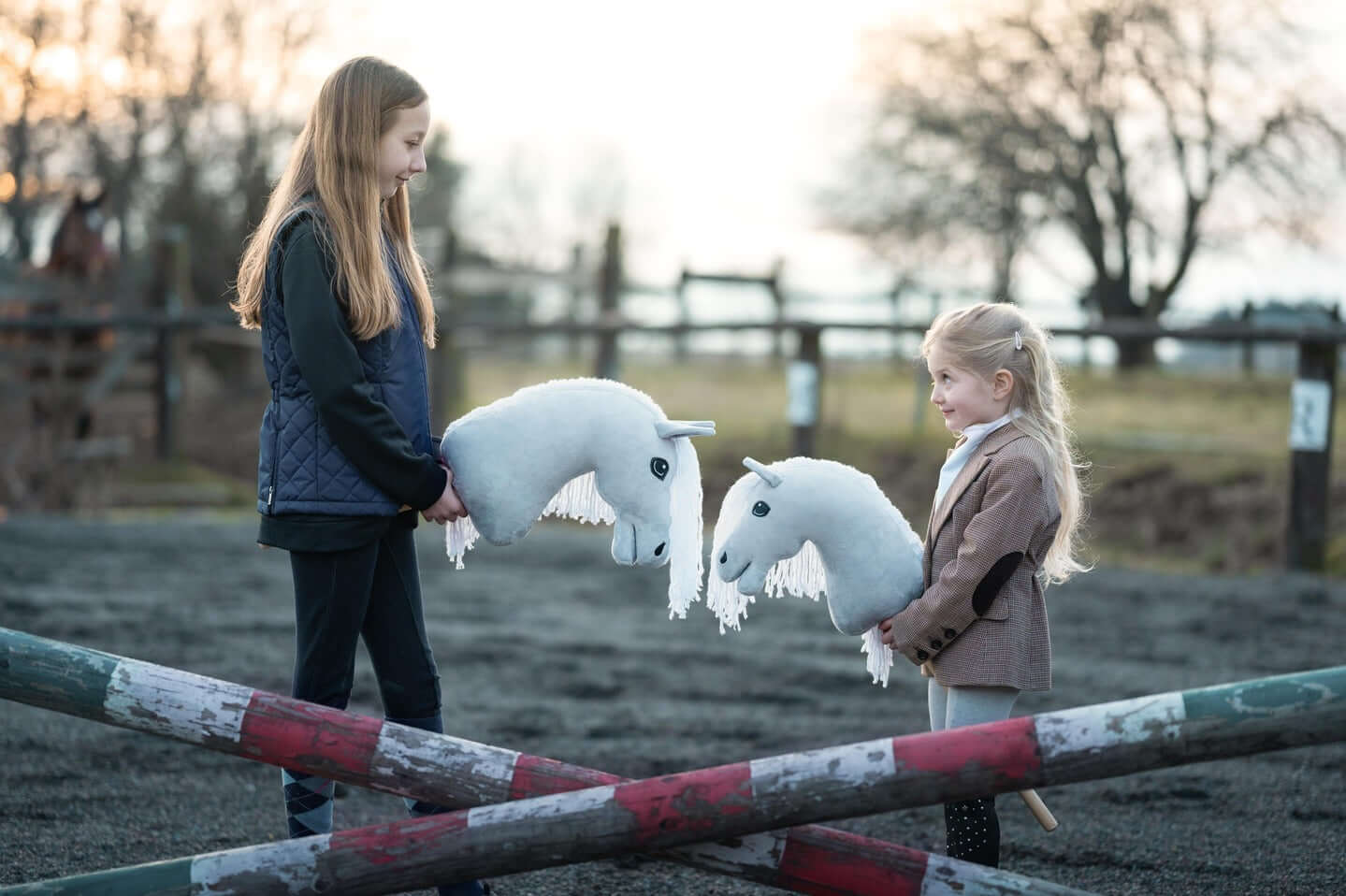 Two children holding white Hobby Horses beside painted jumps in an outdoor arena