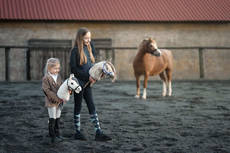 Twee kinderen dragen Hobby Horses in een buitenpiste naast een pony