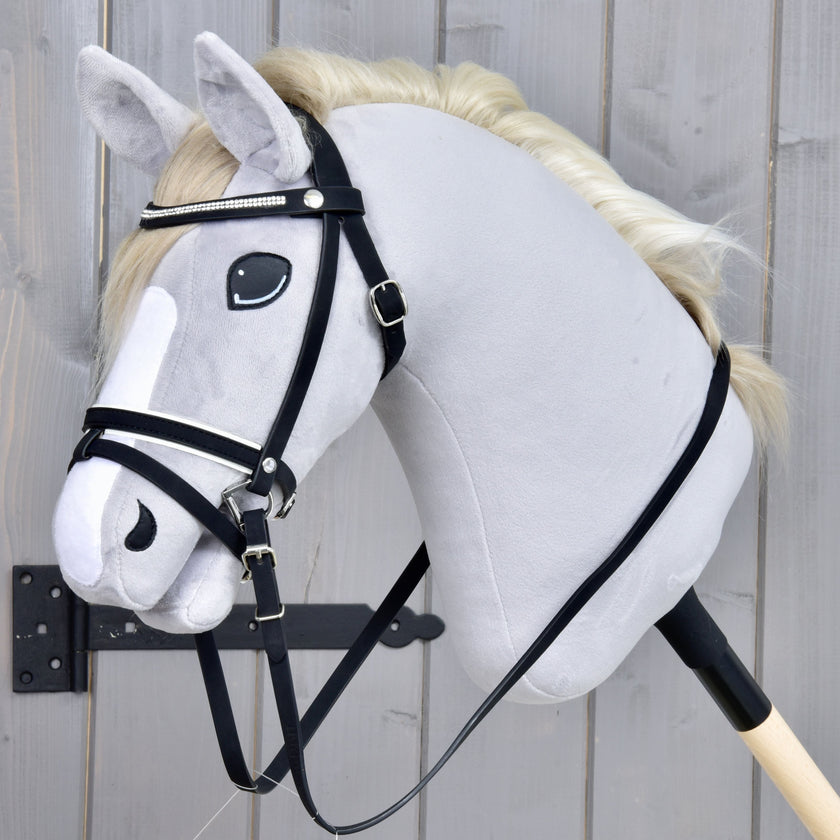 A plush hobby horse with a white mane, black bridle, and a white stripe on its face is attached to a wooden stick, positioned in front of a gray wooden background.