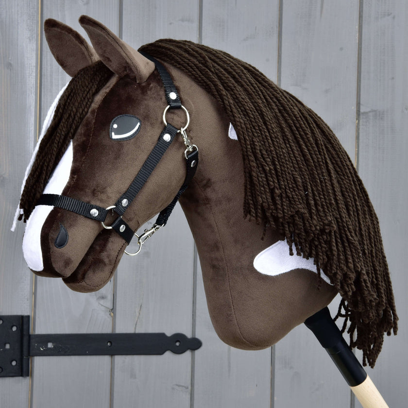 A realistic brown and white hobby horse with a black bridle and long yarn mane, mounted on a wooden stick, is positioned in front of a gray wooden fence.