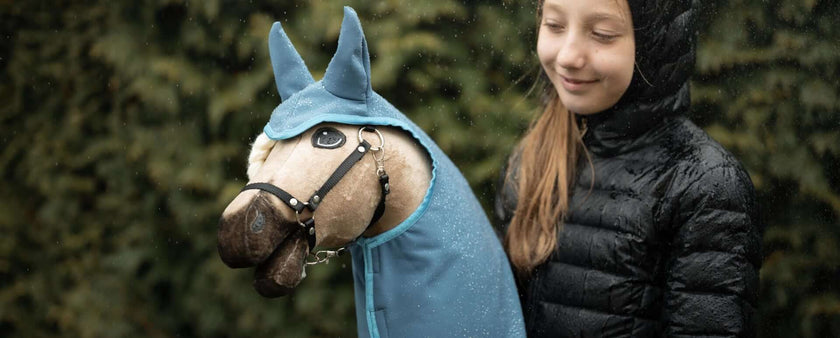 A girl in a black jacket smiles while holding her Hobby Horse, which wears a blue hood and bridle, with greenery softly blurred in the background.