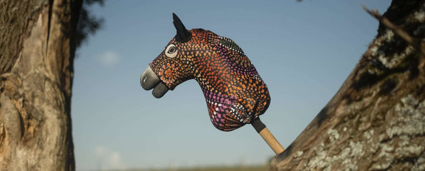 A colorful, patterned hobby horse, perfect for hobby horsing, with black ears and a stick handle is positioned between two tree trunks under a clear blue sky.