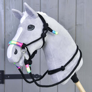 Een grijze pluche hobby horse van Hobby Horse LarDen met een witte bles en witte manen, met het Touwset regenboog-halster, gemonteerd op een houten stok tegen een achtergrond van grijze houten panelen.