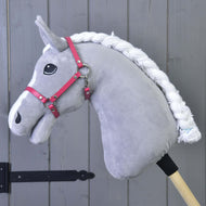 Een grijze pluchen Hobby Horse van Hobby Horse LarDen met witte gevlochten manen, zwart oog en Halster framboos (maat M), bevestigd aan een houten stok, staat voor een grijze houten paneelwand.
