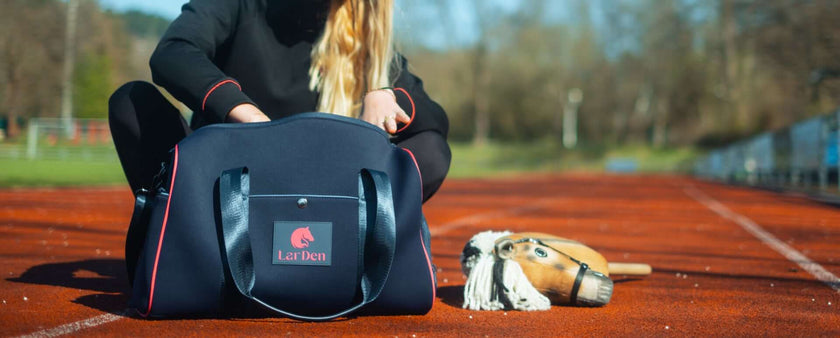 A person with long blonde hair sits on a running track, opening a dark sports bag with LarDen logo. A Hobby Horse toy lies on the ground beside them, hinting at their passion for Hobby Horsing. The background is blurred, showing trees and fencing.