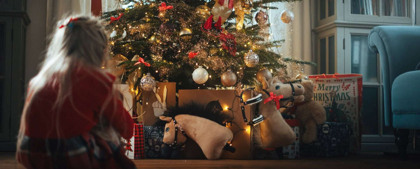 A young child with long blonde hair sits by a decorated Christmas tree, surrounded by gifts, plush rocking horses, and a festive hobby horse, bathed in cozy living room lights.