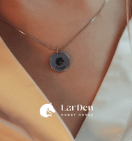 Close-up van een persoon die de Hobby Horse LarDen zilveren ketting draagt met een ronde hanger van 925 sterling zilver, gegraveerd met een paardenkop en "LarDen", met het merklogo en de merknaam, als uiting van passie voor Hobby Horsing.