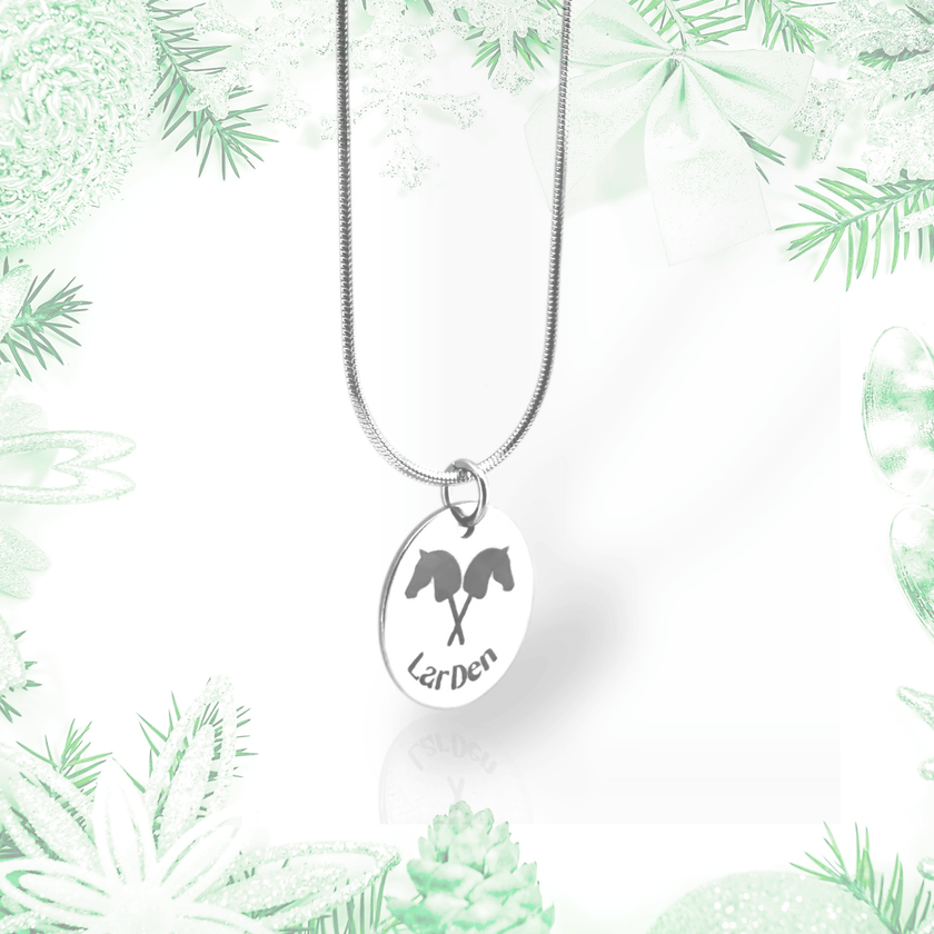 A Necklace with Hobby Horse pendant by Hobby Horse LarDen, crafted from surgical stainless steel, is displayed against a pale green and white festive backdrop with pine branches, bows, and ornaments.