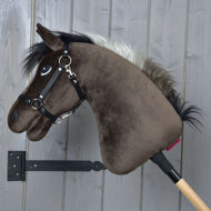 The Hobby Horse LarDen “Hobby Horse Alfredo with Black Halter,” styled like an English Thoroughbred, with a brown head, black mane, and white streak, hangs on a gray wooden wall. The plush stallion features a bridle and embroidered eye.
