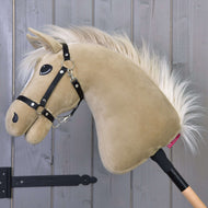 Hobby Horse Dancing Lady met zwart halster van Hobby Horse LarDen heeft een pluchen hoofd van een Engelse volbloedmerrie met een beige vacht en witte manen op een houten stok, weergegeven tegen een grijze houten paneelwand.