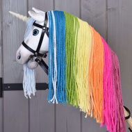 Hobby Horse Eenhoorn Rainbow met Hoofdstel en Halster van Hobby Horse LarDen met een witte hoorn, blauwe ogen, een hoofdstel en levendige regenboogmanen van garen met blauwe, groene, gele, oranje en roze strengen, tegen een houten achtergrond.