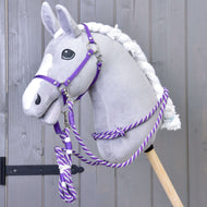 A plush hobby horse with a gray head, white mane, and white blaze features the Hobby Horse LarDen Set Start Purple (halter, cordeo, lead rope, reins), mounted on a wooden stick against a gray wooden wall.