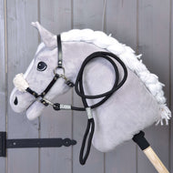 Set Basic van Hobby Horse LarDen toont een pluche Hobby Horse met witte manen, zwart hoofdstel met teugels en zwart halster op een houten stok, leunend tegen grijze houten panelen.
