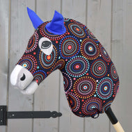 Hobby Horse Beschermpak Alice (Lycra) van Hobby Horse LarDen met een kleurrijk patroon op het hoofd, blauwe vilten oren en expressieve ogen op een houten stok, weergegeven tegen een lichtgrijze houten wand.