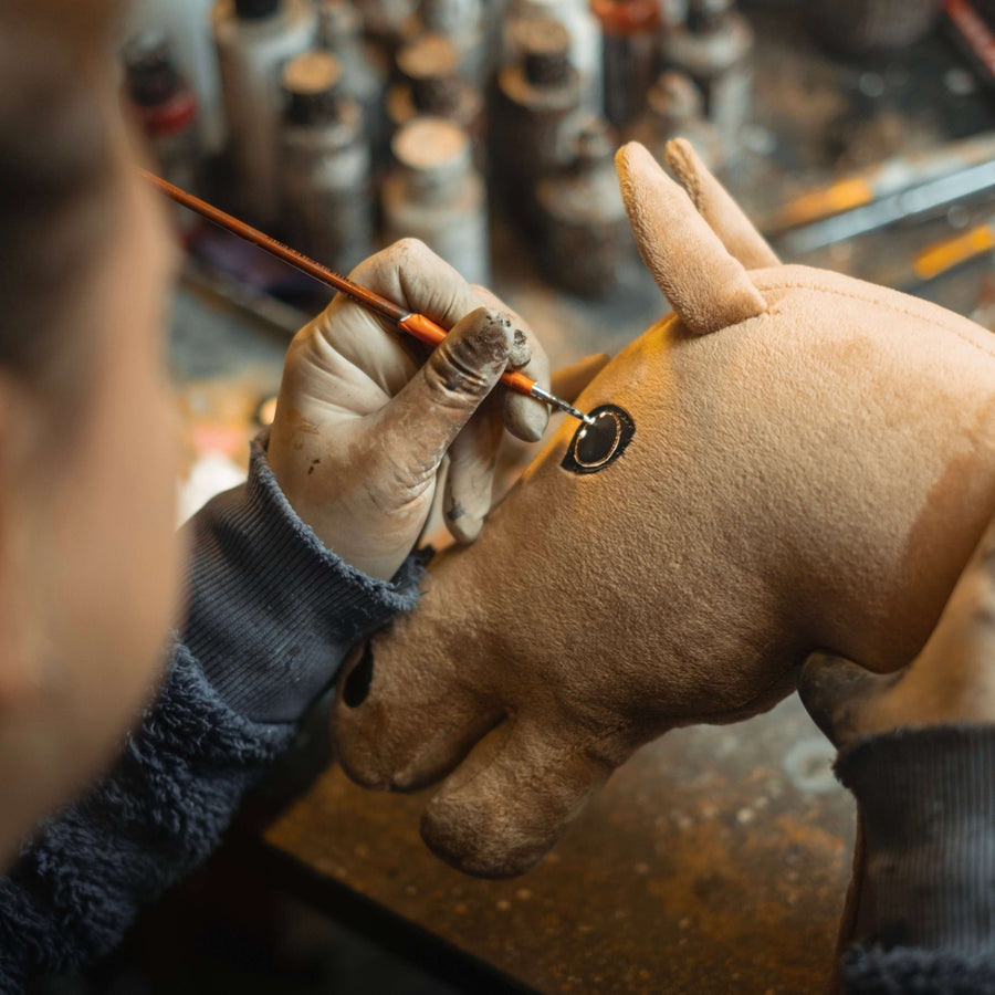A person wearing gloves uses a paintbrush to carefully paint the eye of a brown hobby horse plush animal toy, with jars of paint visible in the blurred background.