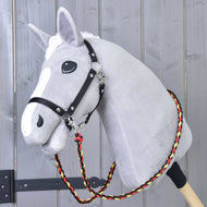 Teugels Flag van Hobby Horse LarDen, een grijze pluche hobby horse op een houten stok met zwart tuig en handgemaakte teugels van gevlochten rood, geel en zwart polypropyleen touw, getoond tegen een houten wand.