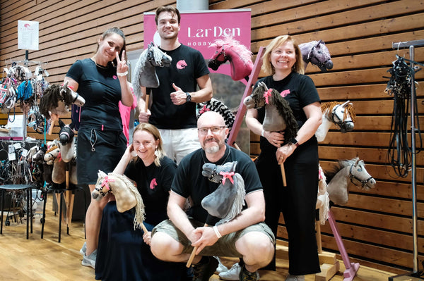 Five smiling adults pose indoors with their hobby horses, celebrating Hobby Horsing. Four stand while one kneels; all wear black shirts. A wooden paneled wall and a pink "LarDen" sign are behind them, with various hobby horses on display.
