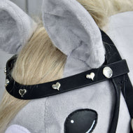 Close-up van een grijze pluchen hobby horse-kop met het Complete De Luxe hoofdstel met 2 frontriemen van Hobby Horse LarDen, met twee afneembare frontriemen versierd met kleine zilverkleurige hartjes en ronde studs. Het paard heeft lichtblonde manen en grote zachte oren.