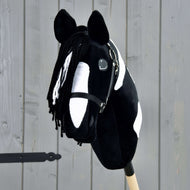 Hobby Horse Merlin met zwart halster van Hobby Horse LarDen, een zwart-wit gevlekte Hobby Horse in de stijl van een Barock Pinto-hengst, met manen van garen en hoofdstel, gemonteerd op een houten stok voor een grijze schutting.