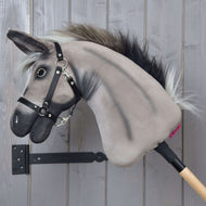 Hobby Horse Ezel Olin met zwart halster van Hobby Horse LarDen met een grijze pluchen ezelkop, donkere manen, een zwart halster voor Hobby Horse, teugels en een zilveren ring bij de mond op een houten stok, tegen een grijze wand.