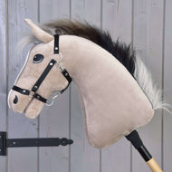Hobby Horse Piccolino met zwart halster van Hobby Horse LarDen, met een lichtbeige gezicht en zwarte manen, is bevestigd op een houten stok en geposeerd voor springreiten voor een grijze houten wand.