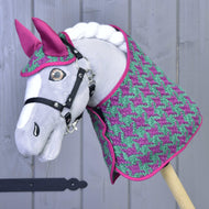 Een grijze Hobby Horse LarDen staat voor een lichtgrijze houten wand en draagt Set Coco: een roze-groene dekenset met pied-de-poulepatroon, een bijpassend roze oornetje en zwart hoofdstel.