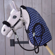 Een pluchen Hobby Horse Set Harmony blauw van Hobby Horse LarDen met zwart halster voor Hobby Horse, teugels en een blauw-zwarte staldeken met pied-de-poulepatroon is aan een wand gemonteerd tegen een lichte achtergrond van houten planken.