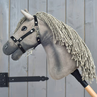 Hobby Horse Swing met zwart halster, maat S van Hobby Horse LarDen, geïnspireerd op de elegantie van een Hannoveraanse hengst, met een grijze manen van garen, een zwart hoofdstel en knoopogen. Het is gemonteerd op een houten stok en gefotografeerd tegen een grijze houten achtergrond.