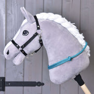 Cordéo Turquoise van Hobby Horse LarDen, een grijs pluchen Hobby Horse met witte manen van garen, zwart hoofdstel, blauwe touwteugels en een handgemaakte turquoise cordeo voor hoofdstelloos rijden, afgebeeld op een stok voor een houten wand.