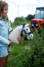 Lifestylefoto: een meisje in een spijkerjasje houdt de Hobby Horse Aurora met zwart Halster van Hobby Horse LarDen naast een heg vast, met een rode tractor vaag op de achtergrond - crèmewitte pluchen kop, zwart-wit gestreepte garenmane - hier getoond met het met bont gevoerde LarDen Blue Halster