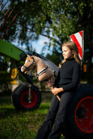 Lifestylefoto: een meisje in een zwarte coltrui staat naast groene landbouwmachines en houdt de Hobby Horse Bren met zwart Halster van Hobby Horse LarDen vast - zandbruine pluchen kop, crèmekleurige garenmane - hier getoond met het Espresso-borsttuig met bont