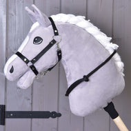 Cordéo zwart van Hobby Horse LarDen, een grijs pluchen Hobby Horse met witte manen op een houten stok, met een zwart hoofdstel, teugels en handgemaakte cordeo, afgebeeld tegen een achtergrond van een grijze houten wand.