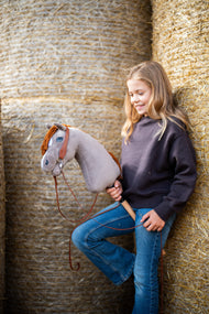 Lifestylefoto: een glimlachend meisje in een donkerbruine sweater leunt tegen ronde strobalen en houdt de Hobby Horse Chanell met zwart Halster van Hobby Horse LarDen vast - grijze pluchen kop, blauw oog, rosse garenmane - hier getoond met het volledige Western-hoofdstel met één oorstuk