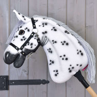 Hobby Horse Chantal met zwart halster van Hobby Horse LarDen met witte vacht met zwarte vlekken, grijze manen van garen, zwart halster en gemonteerd op een houten stok tegen een lichtgrijze houten wand