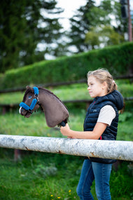 Lifestylefoto: een meisje in een donkerblauw gewatteerd gilet staat achter een witte paddockreling en houdt de Hobby Horse Chianti met zwart Halster van Hobby Horse LarDen vast, met een geschoren groene heg erachter - donkerbruine pluchen kop, donkere garenmane - hier getoond met het met bont gevoerde Teddy Petrol Halster