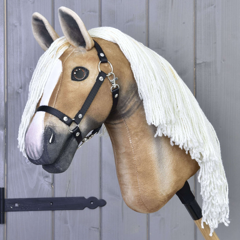 The Hobby Horse LarDen “Connie with Black Halter” features a realistic tan and white design, a long white yarn mane, and is shown mounted on a stick against a gray wooden wall.