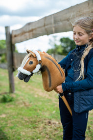 Lifestylefoto: een meisje in een donkerblauw gilet staat bij een houten paddockomheining en houdt de Hobby Horse Connie met zwart Halster van Hobby Horse LarDen vast - voskleurige pluchen kop met een donkere aalstreep, crèmekleurige garenmane - hier getoond met het met bont gevoerde Teddy Classic Halster