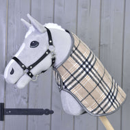 De Hobby Horse LarDen "Deken Cremo England" is een grijze Hobby Horse met een zwart halster voor Hobby Horse en een beige geruite deken, met een oornetje voor bescherming tegen insecten, gemonteerd op een houten stok tegen een grijze houten wand.