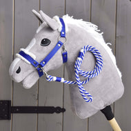 Set Essential Blauw van Hobby Horse LarDen op een grijze pluche Hobby Horse met witte manen, blauw halster met bontrand en blauw-wit touwteugel op een houten stok, getoond voor een grijze houten schutting.