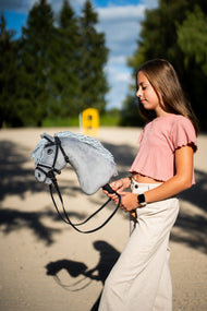 Lifestylefoto: een meisje in een roze top loopt met de Hobby Horse Grace met zwart Halster van Hobby Horse LarDen over een zandpiste - grijze pluchen kop, lichtgrijze garenmane - hier getoond met het volledige De Luxe hoofdstel met twee frontriemen