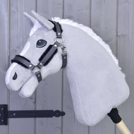 Halster met bont - kunstleer Graphite Volledig van Hobby Horse LarDen is gemonteerd op een houten stok en leunt tegen een lichtgrijze houten wand, met een grijs pluchen hoofd, witte manen, zwart hoofdstel en halster van kunstleer.