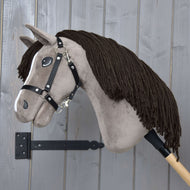 Hobby Horse Guccini met Zwart Halster van Hobby Horse LarDen met bruine manen van garen en een houten stok, tegen een grijze wand, ideaal voor jonge fans van de paardensport.