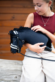 Lifestylefoto: een meisje in een bordeauxrode top houdt Hobby Horse Mahal met zwart Halster van Hobby Horse LarDen in haar armen voor een houten wand – zwarte pluchen kop, zwarte manen van garen, witte bontneusriem – hier getoond met het complete hoofdstel met bont
