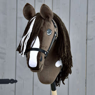 Handgemaakte Hobby Horse Monte met zwart halster van Hobby Horse LarDen, met een bruin-witte vacht geïnspireerd op het American Paint Horse, een zwart halster, lange manen van garen en gemonteerd op een houten stok tegen een grijze houten achtergrond.