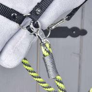 Close-up van een metalen sluiting die een zwarte band bevestigt aan de geel, zwart en grijze Hobby Horse - Teugels citroen streep van Hobby Horse LarDen. De achtergrond is onscherp grijs hout met een deel van een zacht wit object zichtbaar.