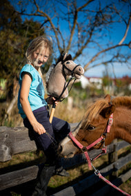 Blond meisje in een turquoise top op een houten paddockhek met Hobby Horse Piccolino met zwart Halster van Hobby Horse LarDen, lichtbeige pluchen kop met donkere garenmane, met een voskleurige pony achter haar en het Complete zwarte hoofdstel van kunstleer
