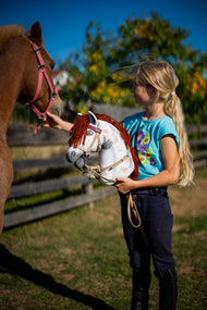 Meisje in een turquoise T-shirt met Hobby Horse Rufus met zwart Halster van Hobby Horse LarDen, een witte Appaloosa-gevlekte pluchen kop met rode garenmane, naast een voskleurige pony in een zonnige paddock, met de kunstleren set Bordeaux met hoofdstel, teugels en martingaal