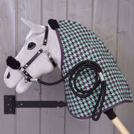 Set Harmony Groen van Hobby Horse LarDen: een pluche Hobby Horse met een zwart halster voor Hobby Horse, groene en paarse staldeken met pied-de-poulepatroon en opgerold zwart touw, gemonteerd op een houten muurbeugel tegen een grijze houten wand.