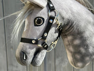 Close-up van Hobby Horse Untamed Sweety van Hobby Horse LarDen met de grijze merriekop, een zwart halster en een realistisch glazen oog, tegen een houten achtergrond.